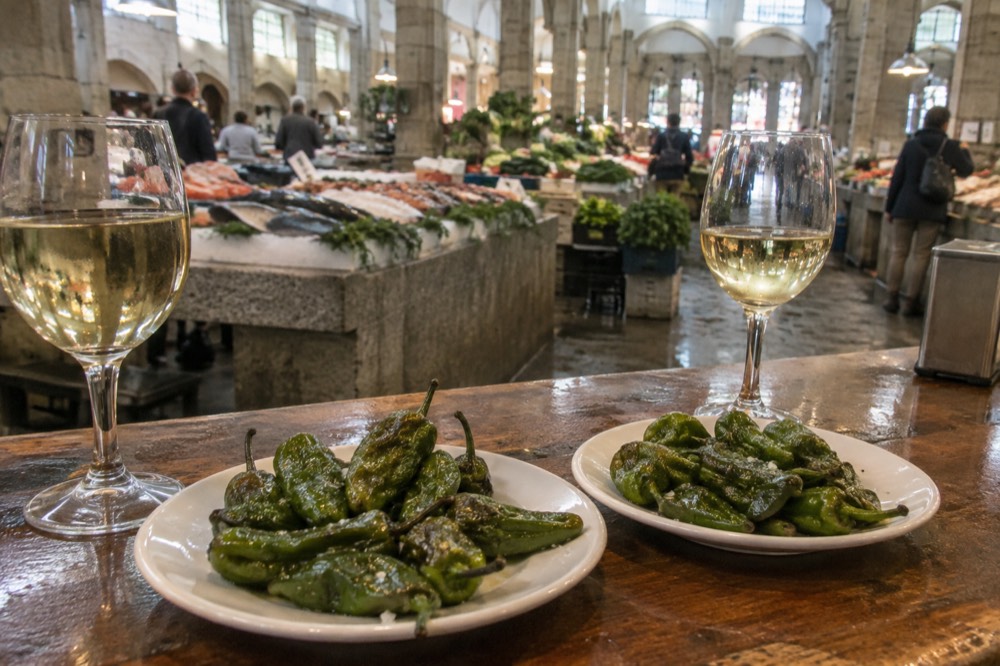 Two plates of fried green peppers on a wooden bar counter beside glasses of white wine