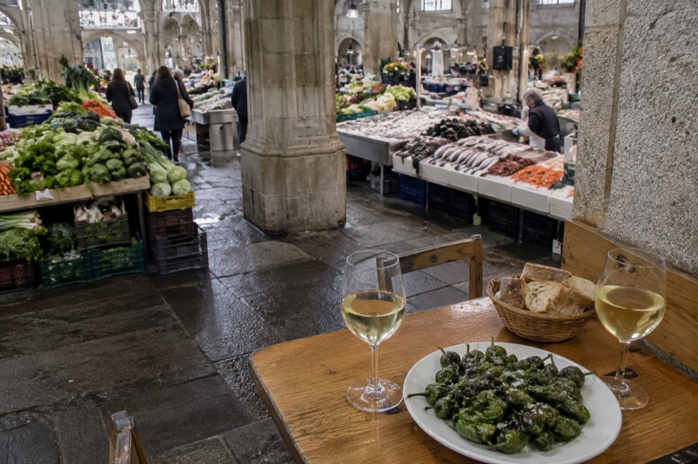 A wooden table set with a plate of green peppers, glasses of white wine and a view of a Galician stone arcade