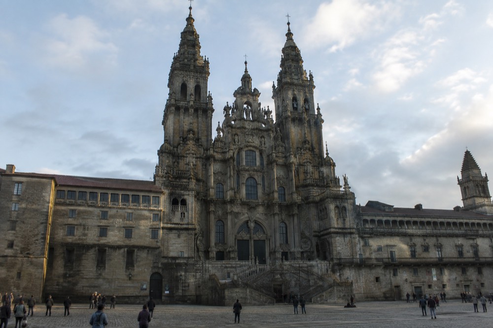 Candlelit side chapel in Santiago de Compostela Cathedral, flickering votive candles and worn stone arches, figures small and distant near the entrance