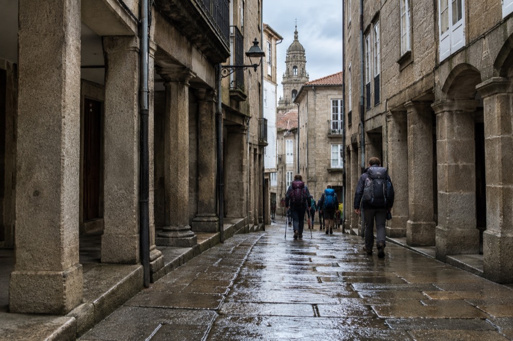A narrow granite street in the old town of Santiago de Compostela with pilgrims walking downhill carrying backpacks