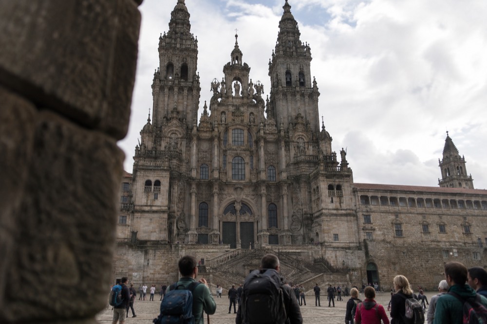 Pilgrims with backpacks standing in a granite square looking up at a baroque cathedral facade in Santiago de Compostela