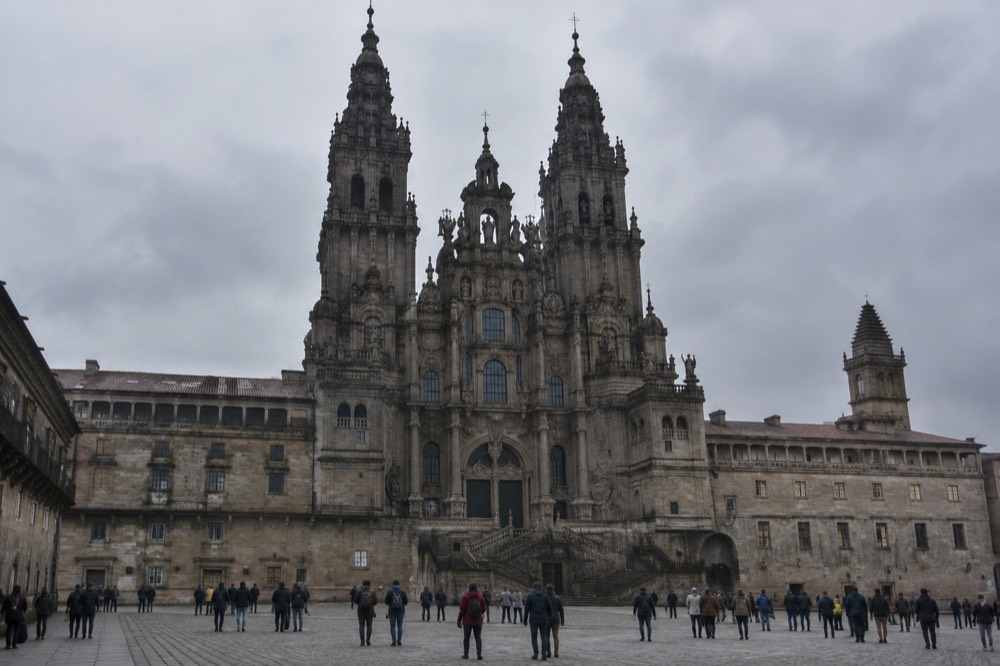 The interior of a granite church in Santiago de Compostela with rows of seats and pilgrims seated for a midday service