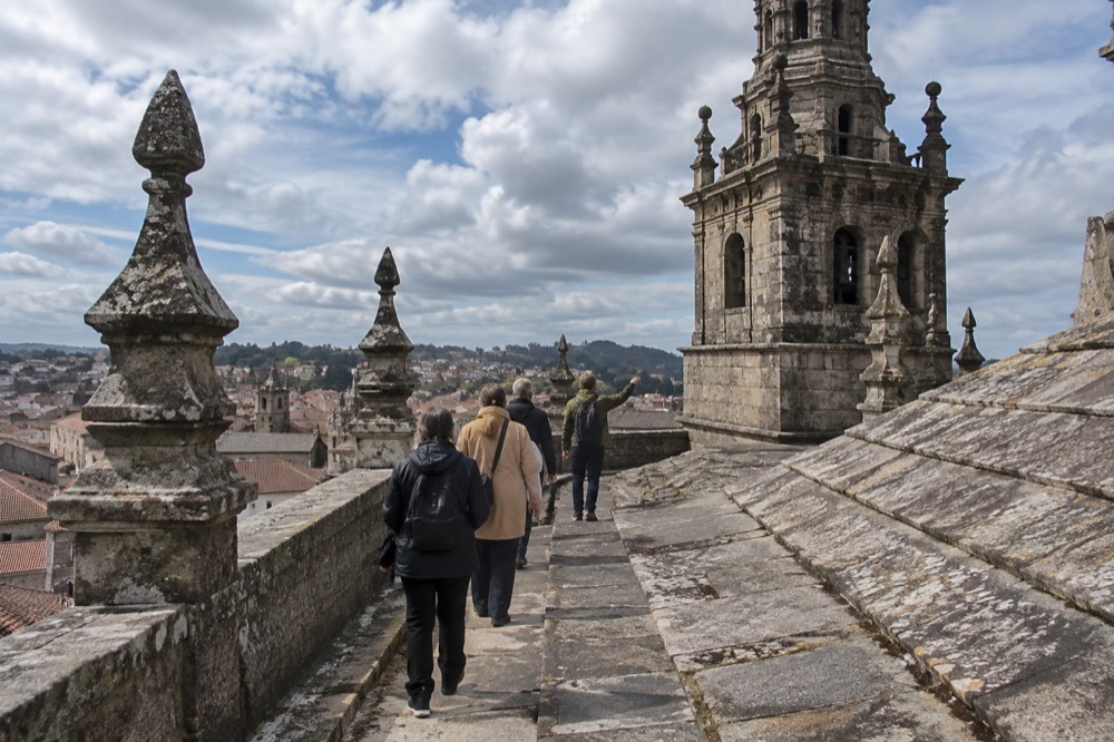 A guide leading a small group of visitors along a cathedral rooftop walkway above the granite rooftops of Santiago de Compostela