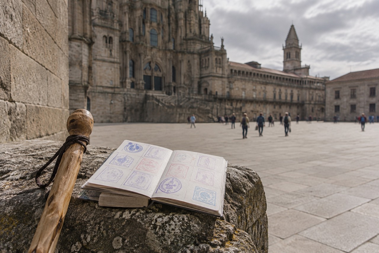 A pilgrim passport booklet held open on a stone wall beside a walking staff, its pages filled with rectangular ink stamps