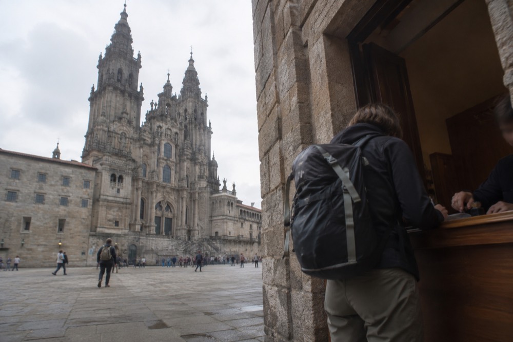 A pilgrim reaching a wooden counter in a stone building to have a rubber stamp pressed into an open booklet