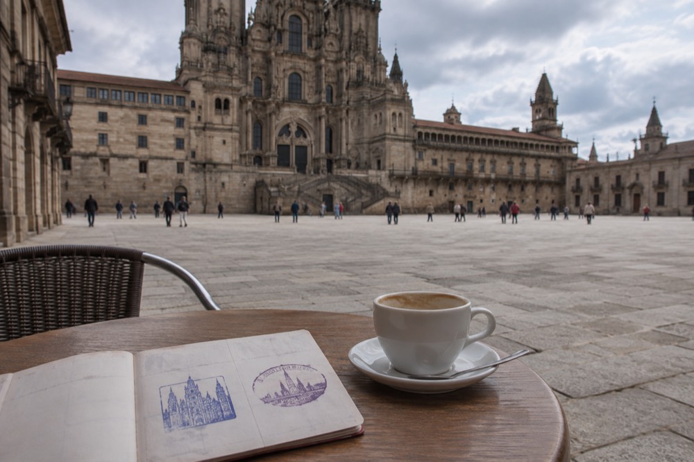 A credencial open on a cafe table next to a coffee cup, showing a day's two ink stamps with dates and place names
