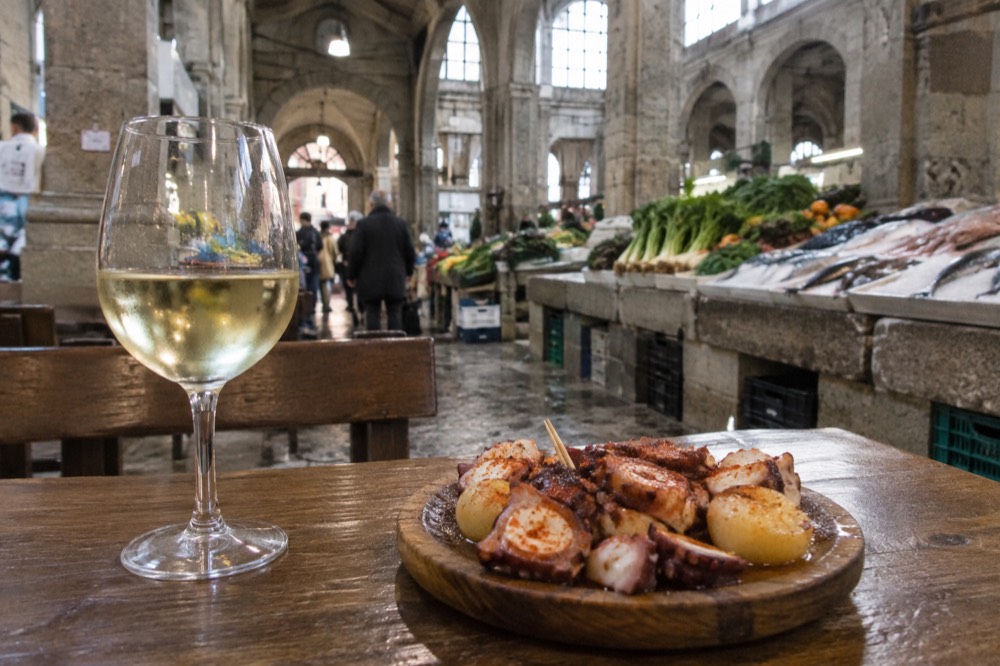 A glass of white wine beside a wooden plate of octopus and potatoes on a tavern table