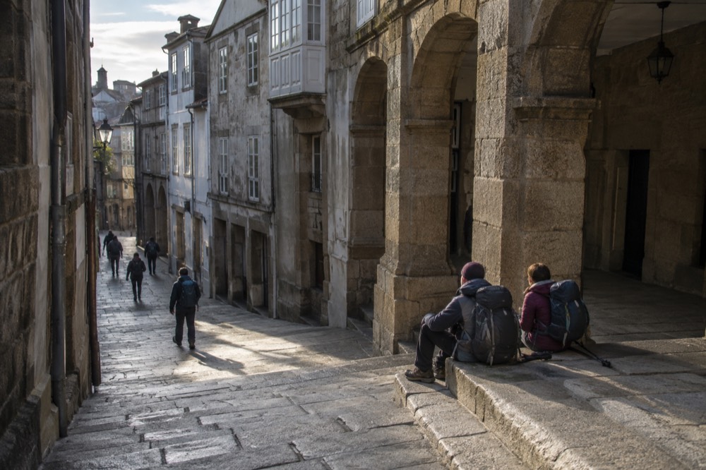 Pilgrims with backpacks resting on stone steps near a granite arcade in the old town