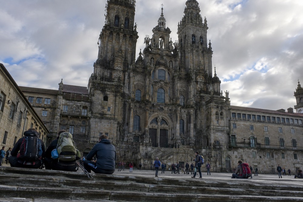 Pilgrims with large backpacks resting on the stone steps of a square in Santiago de Compostela