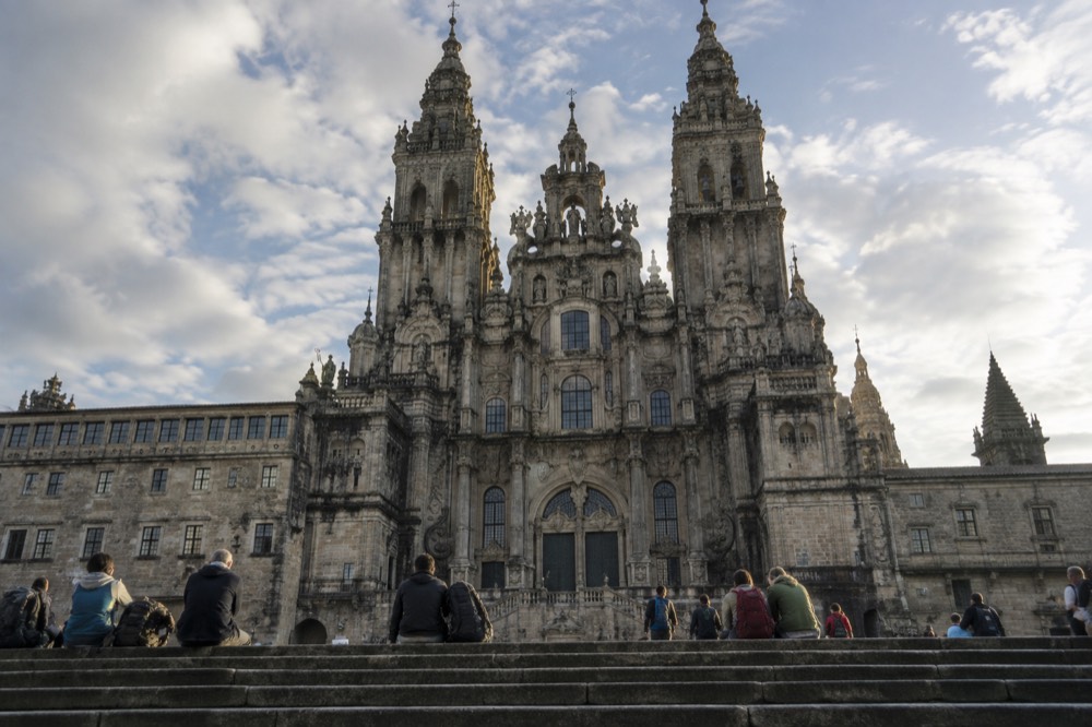 Tired smiling pilgrims with backpacks resting on the stone steps in front of Santiago de Compostela Cathedral