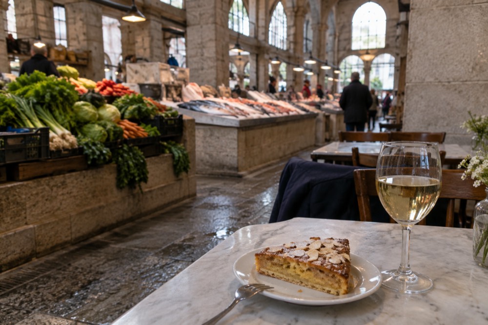 A slice of almond cake on a plate beside a small glass of white wine on a restaurant table with stone walls