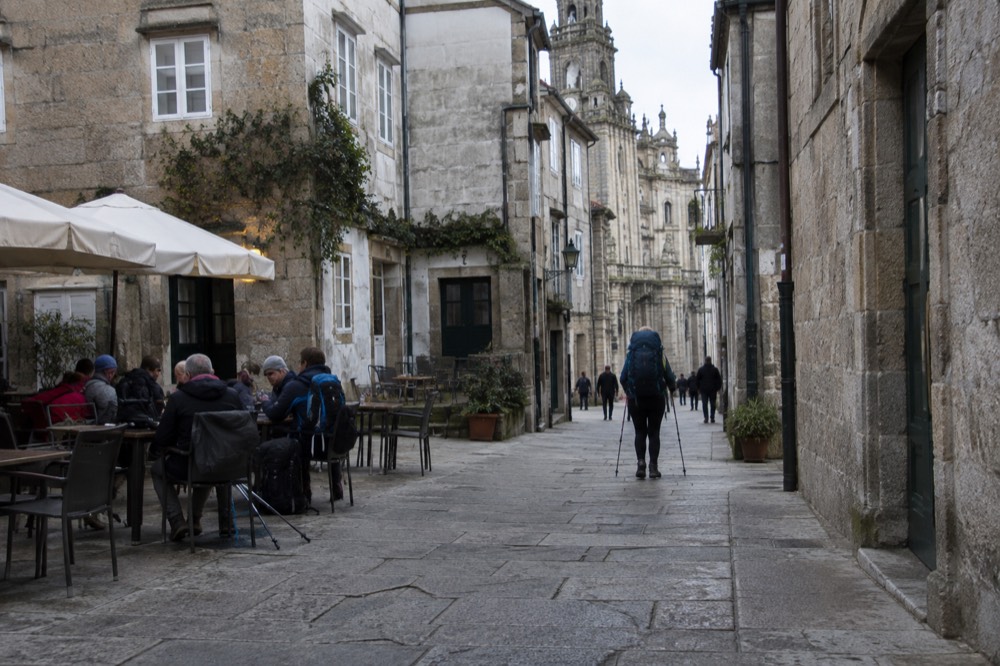 Pilgrims with backpacks and walking poles sitting together at an outdoor table in a small square