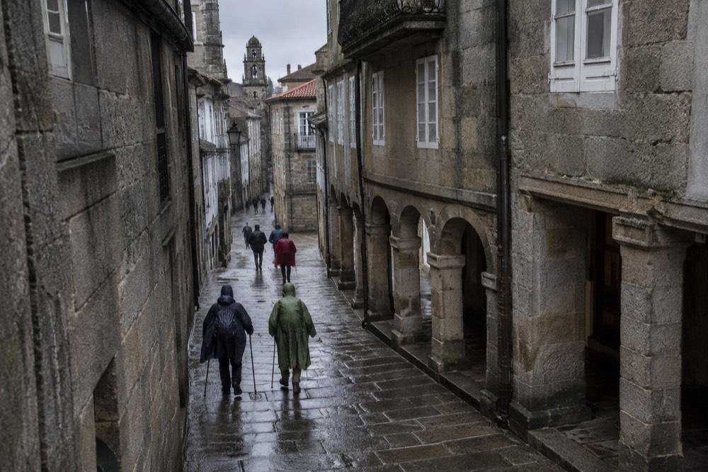Pilgrims in wet rain capes walking into the old town past stone buildings