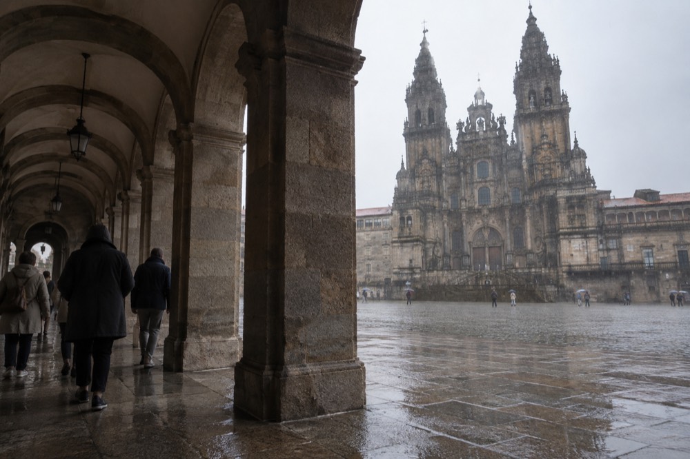 A covered stone arcade with people walking under it while rain falls on the open square beyond