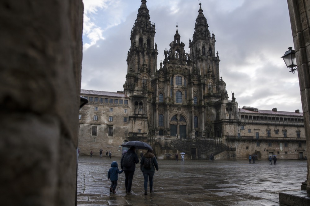 A family in rain jackets walking under an umbrella across a wet stone square in Santiago de Compostela